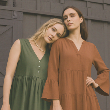 Two women in green and brown dresses standing against a wooden wall.