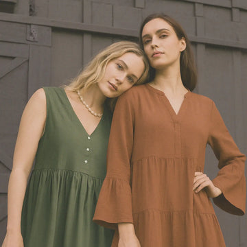 Two women wearing green and brown dresses standing against a wooden wall.